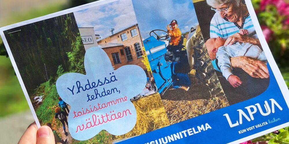 Henkilön käsi pitelee Lapuan kaupungin hyvinvointisuunnitelman 2026–2029 paperista esitettä. Esitteen kannessa on useita kuvia, kuten lapsi traktorilla ja ikäihminen vauvan kanssa. Kansilehden tekstissä lukee ”Yhdessä tehden, toisistamme välittäen” ja ”LAAJA HYVINVOINTISUUNNITELMA 2026–2029”. Kuva on otettu aurinkoisena päivänä, ja taustalla näkyy kukkia ja viheralue.